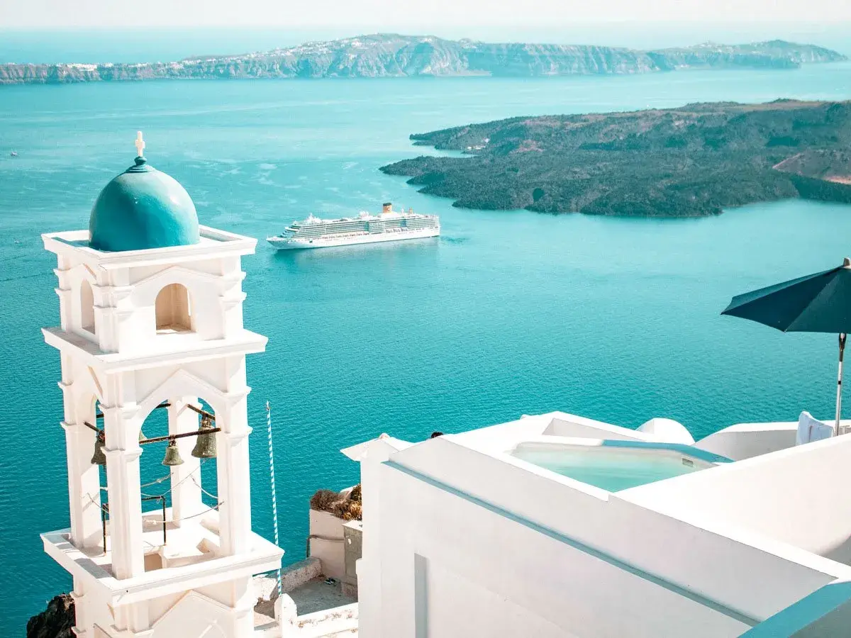 Traumhafter Urlaub Griechenland: Weißer Glockenturm mit blauer Kuppel, Kreuzfahrtschiff auf dem Meer, Infinity-Pool und Sonnenschirm.