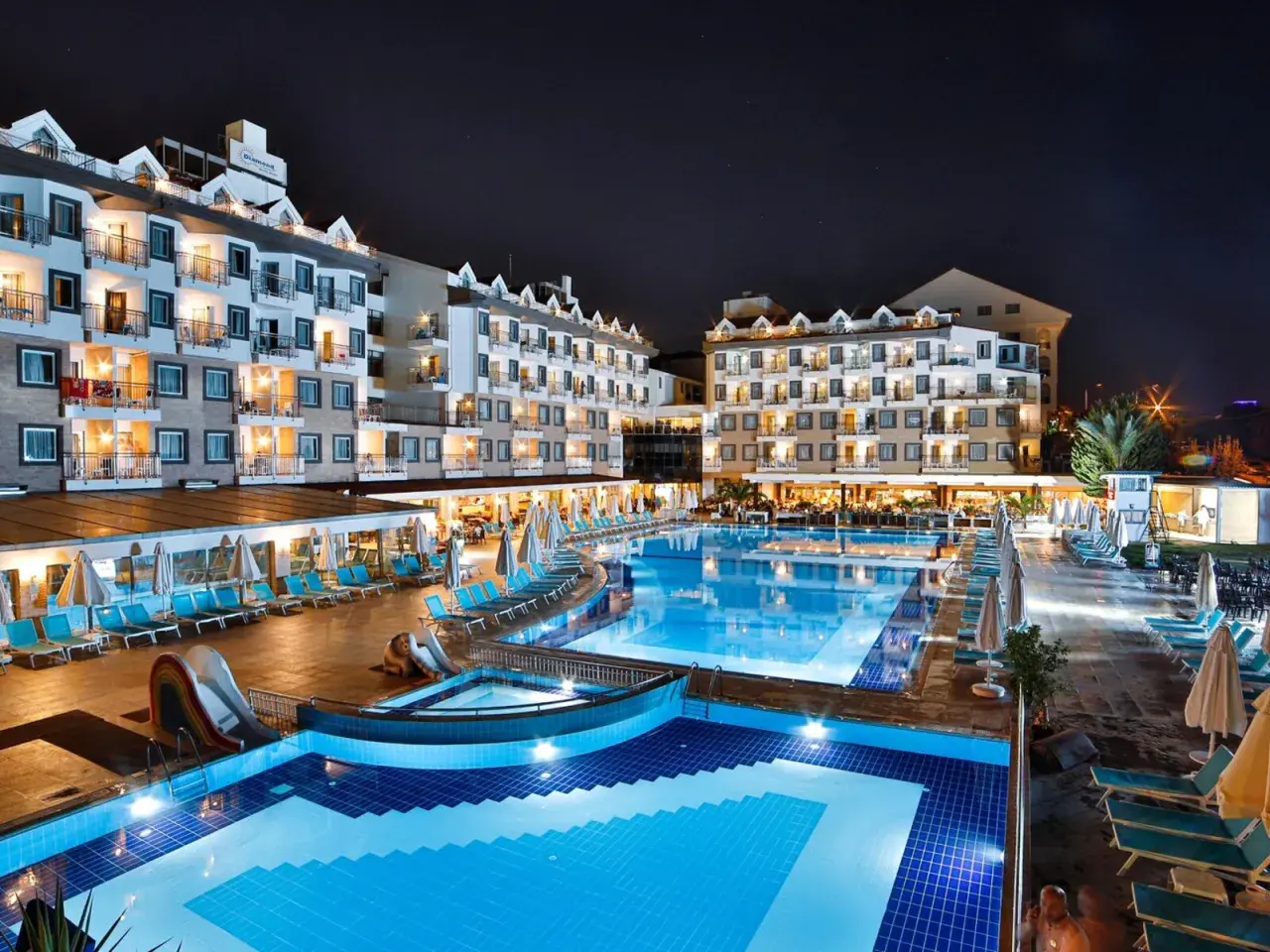 Nachtansicht des Diamond Beach Hotel & Spa mit beleuchteten Pools, Rutschen und Liegestühlen.