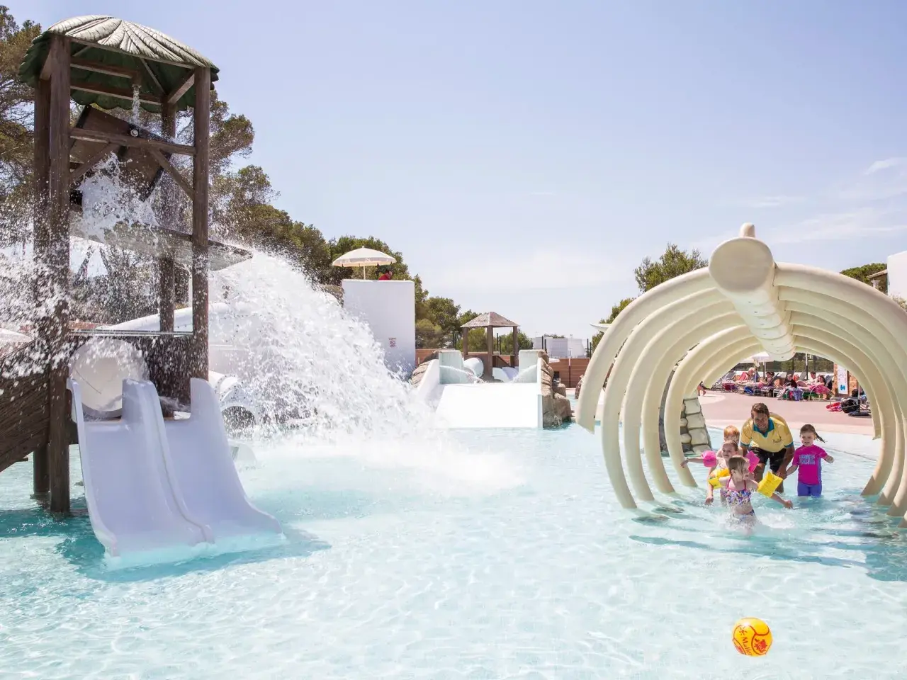 Kinder planschen im Wasserpark, während ein Wasserfall vom Rutschenturm herabstürzt. Ein Mann mit gelbem Shirt beaufsichtigt die Kleinen unter einem knochenartigen Tunnel in den Calimera Fido Gardens.