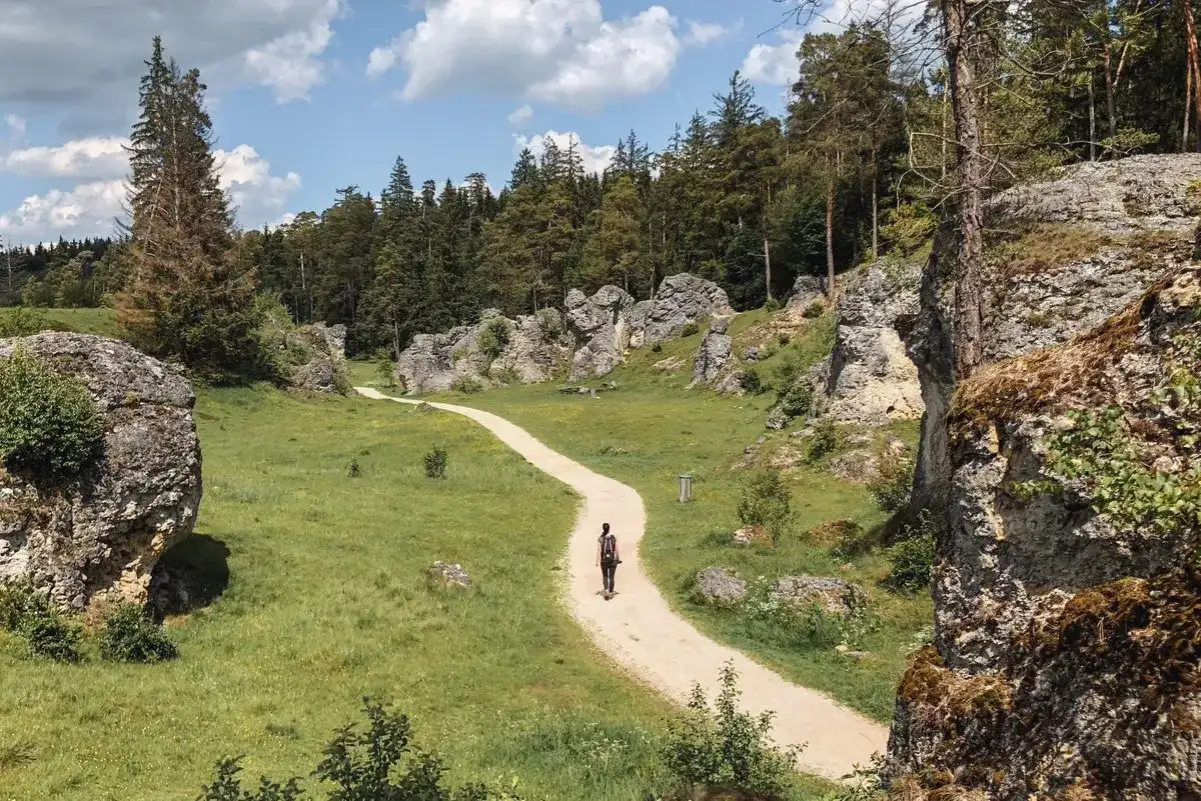 Frau wandert auf einem Kiesweg durch eine felsige Landschaft mit grünen Wiesen und Bäumen. Perfekt für einen entspannten outdoor urlaub.