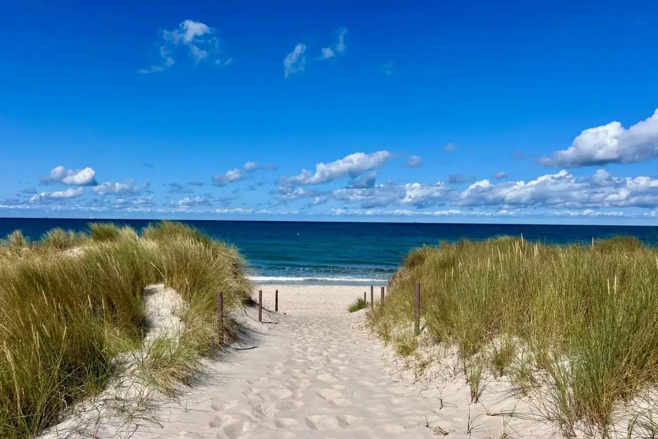 Sandiger Weg durch Dünen zum blauen Meer. Perfekter Urlaub am Meer mit Sonnenschein und Wolken.