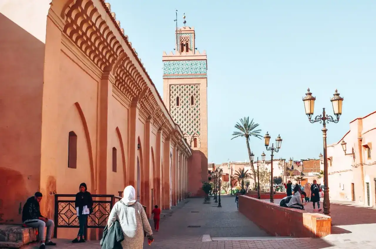 Ein Urlaub in Marrakesch: Ein Minarett ragt über eine Straße mit Palmen und Laternen. Menschen spazieren entlang der rosafarbenen Mauern.