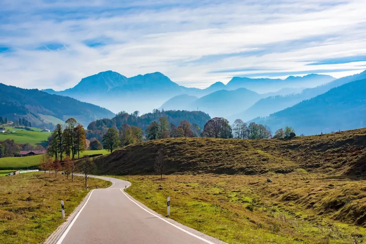 Kurztrip-Ideen mit Auto: Eine kurvige Straße führt durch eine malerische Alpenlandschaft mit sanften Hügeln, bunten Bäumen und blauen Bergketten im Hintergrund.