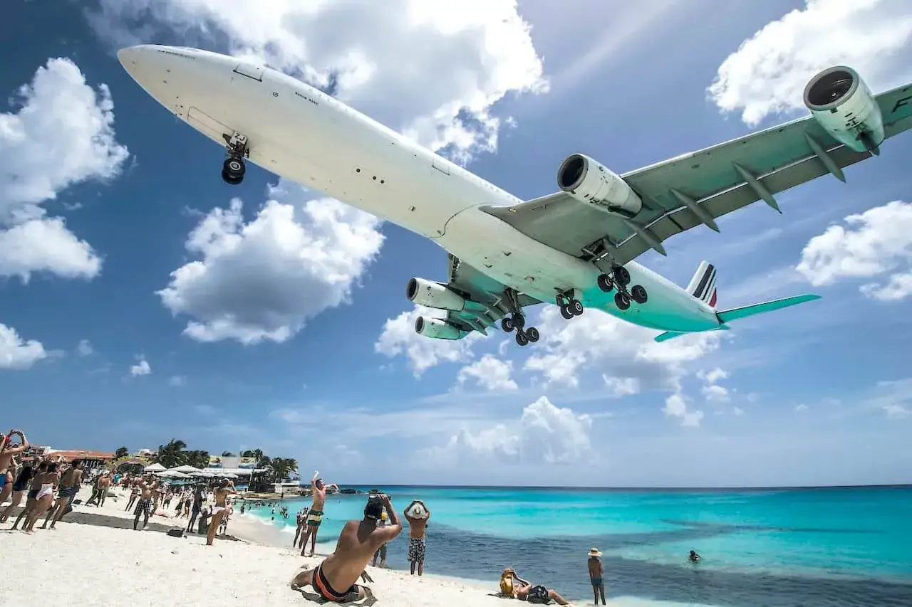 Ein Flugzeug landet tief über einem belebten Strand. Urlauber blicken fasziniert nach oben. Ein Flughafen direkt am Strand, ein unvergesslicher Anblick.