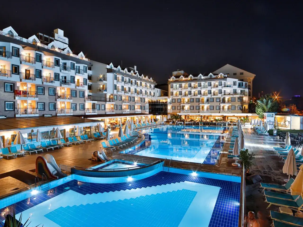 Nachtansicht des Diamond Beach Hotel & Spa mit beleuchteten Pools, Rutschen und Liegestühlen.