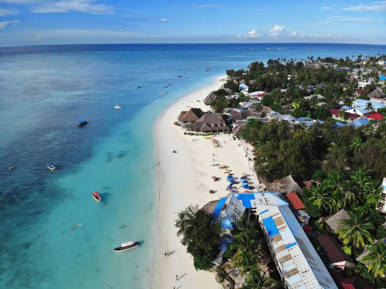 Traumhafter Zanzibar Urlaub: Türkises Meer, weißer Sandstrand und palmengesäumte Bungalows laden zum Entspannen ein. Boote schaukeln im Wasser.