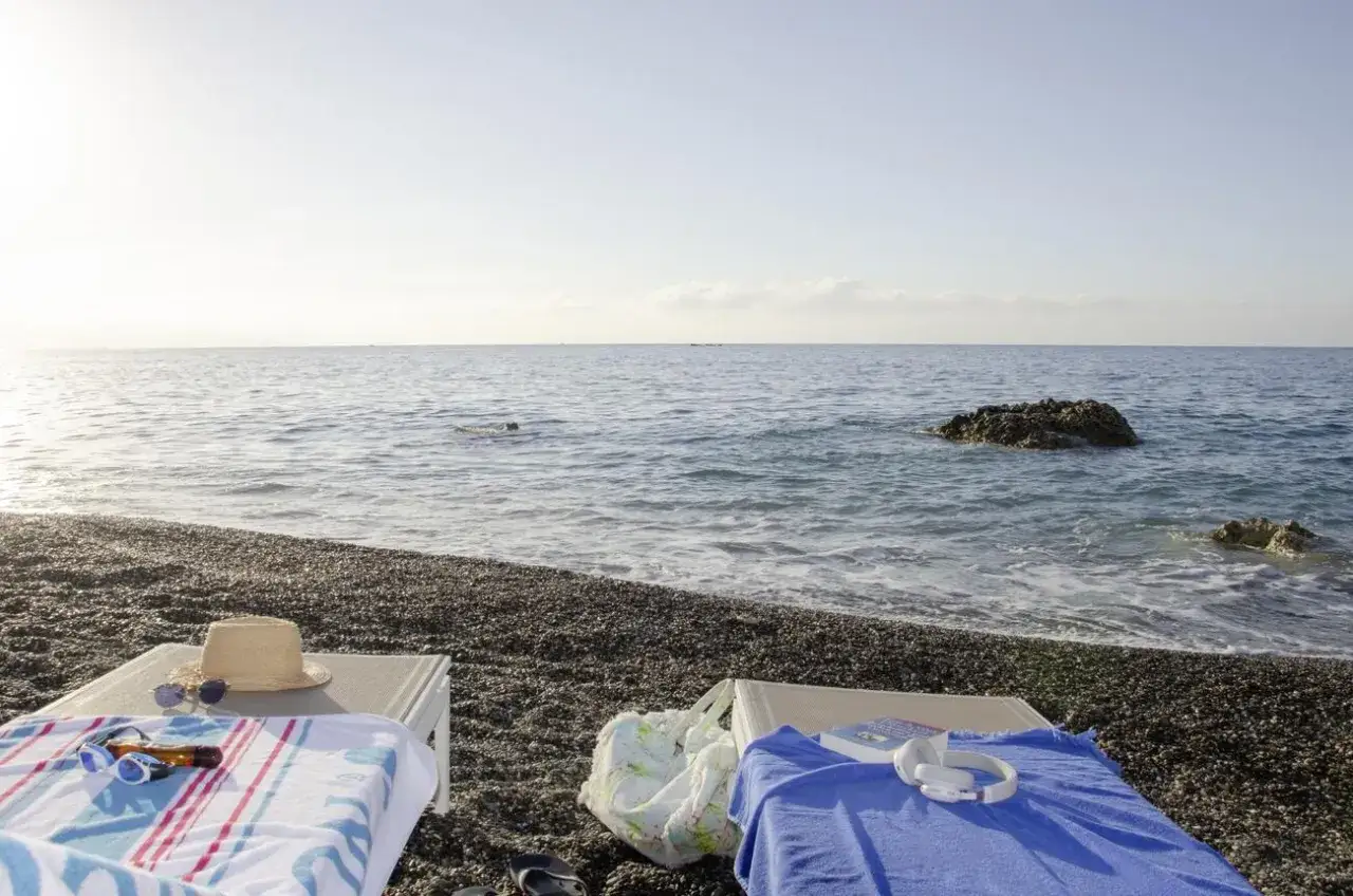 Zwei Liegen am Strand von Robinson Ierapetra, bereit f&uuml;r einen entspannten Tag am Meer mit Hut, Sonnenbrille und Buch.