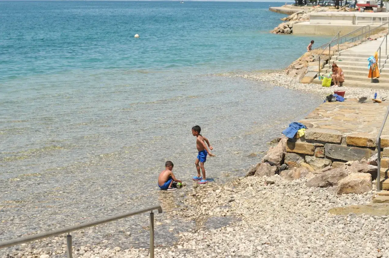 Zwei Jungen spielen am flachen, steinigen slowenien strand. Das klare Wasser lädt zum Planschen ein, während im Hintergrund eine Frau auf den Stufen sitzt.