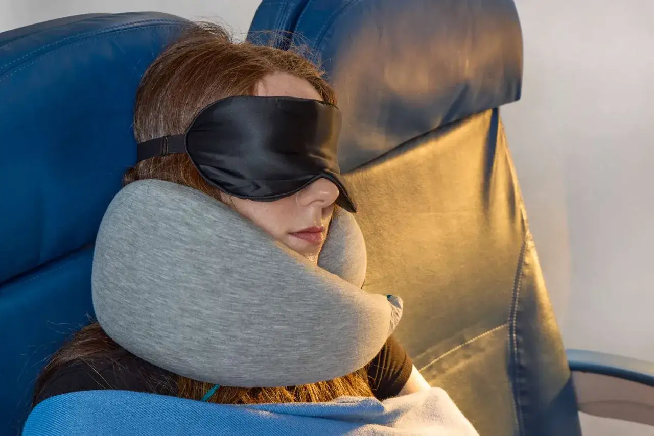 Frau schläft im Flugzeug mit Schlafmaske und Nackenkissen. Ein Nackenkissen im Flugzeug ist sinnvoll für entspanntes Reisen.