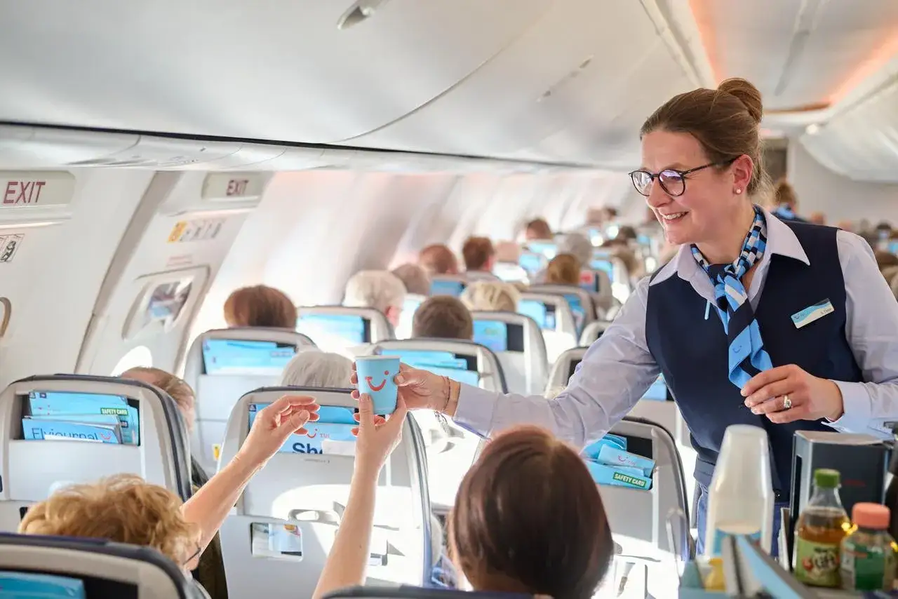 Flugbegleiterin reicht lächelnd ein Getränk an Bord. Bei TUI fly ist die Sitzplatzreservierung kostenlos, was den Flugkomfort erhöht.