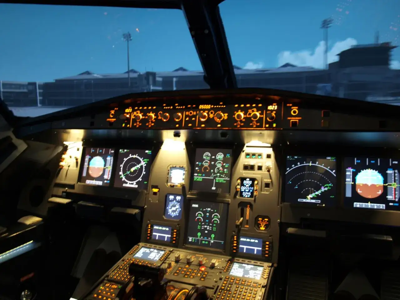 Blick ins Cockpit: Anzeigen leuchten, bereit zum im Cockpit mitfliegen. Draußen dämmert es, Gebäude und Lichter sind zu sehen.