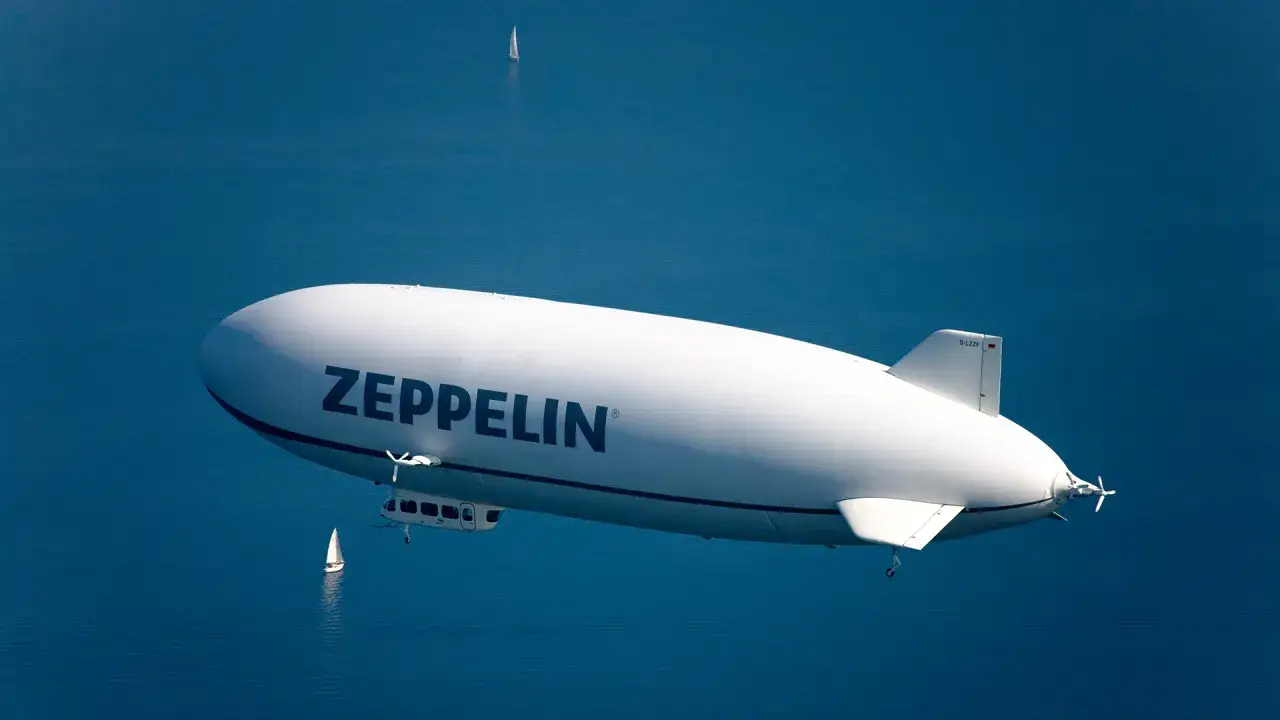 Ein gro&szlig;er wei&szlig;er Zeppelin schwebt &uuml;ber blauem Wasser. Daneben segelt ein kleines Segelboot. Man fragt sich, wie viele Zeppeline es wohl gibt.