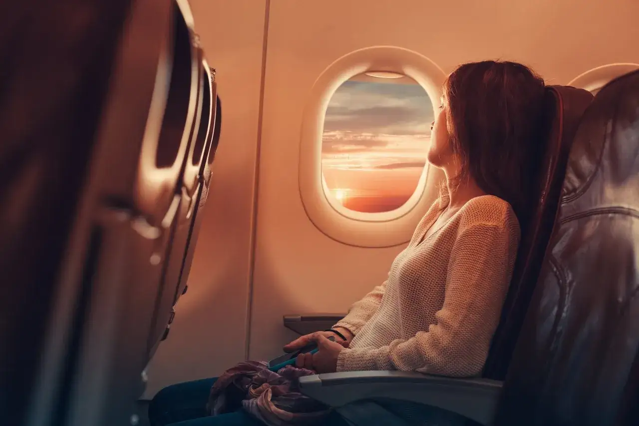 Frau blickt aus dem Fenster im flugzeug, während die Sonne untergeht und den Himmel in warme Farben taucht.