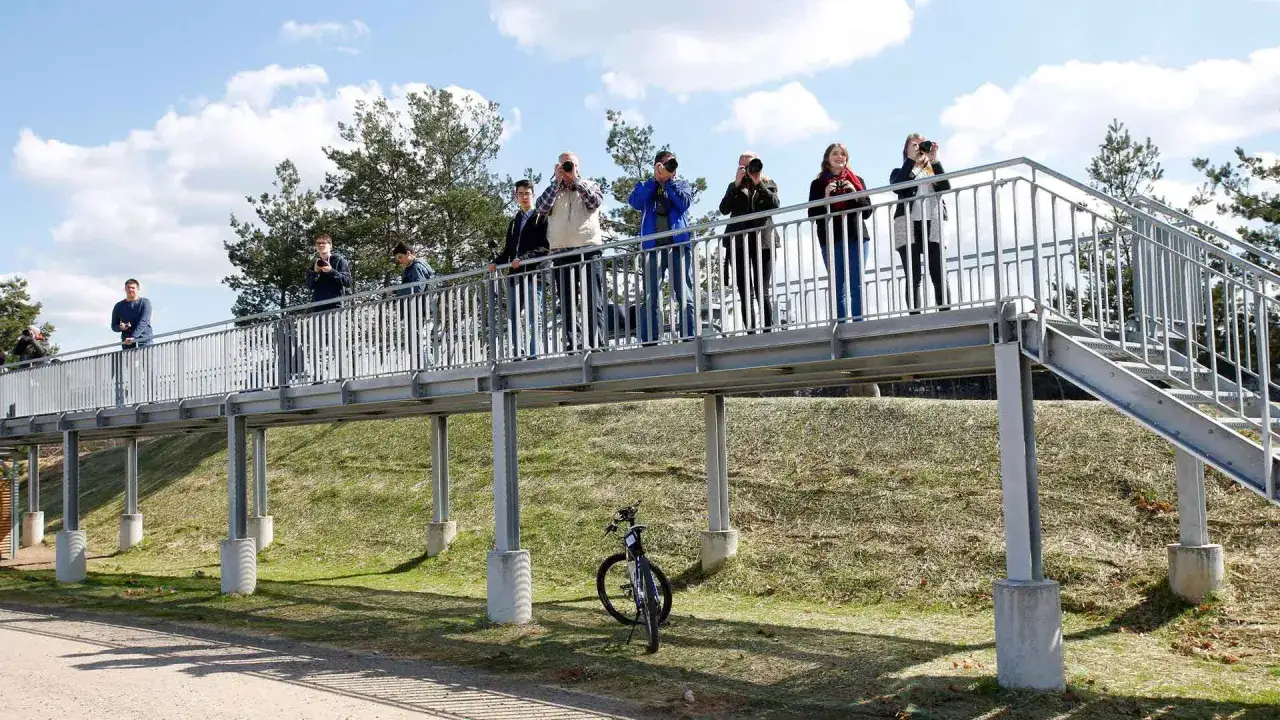 Menschen stehen auf einer Br&uuml;cke und machen Fotos, ein Hobby wie plane spotting. Ein Fahrrad steht im Gras.