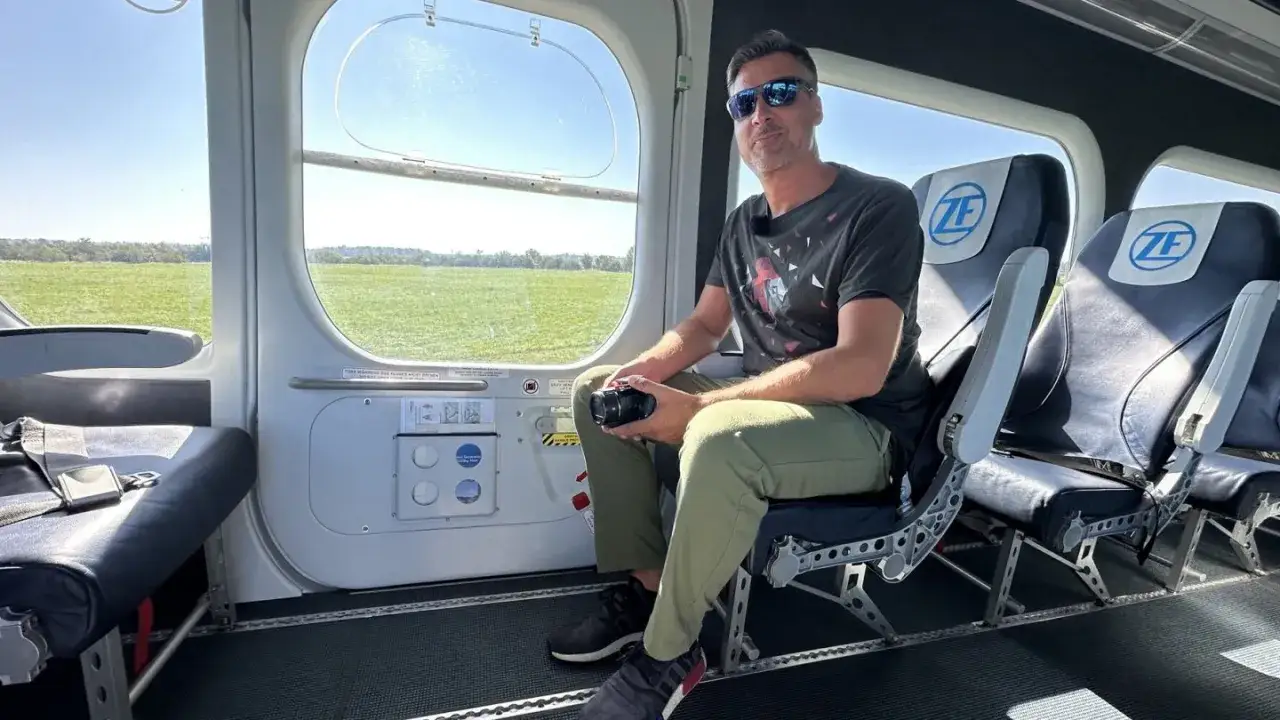 Mann mit Kamera sitzt im Zeppelin innen, blickt auf grüne Felder.