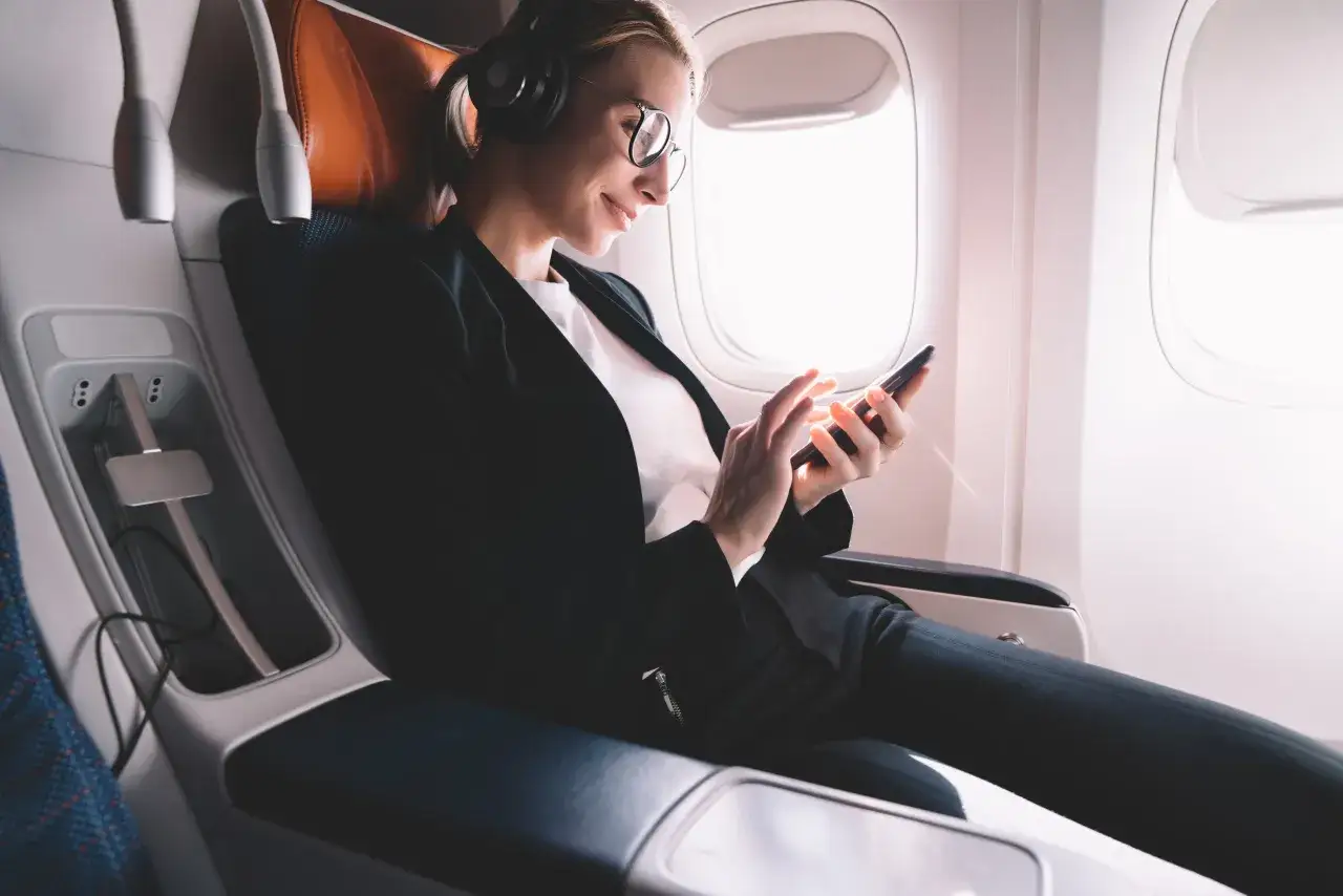 Frau mit Kopfhörern und Brille nutzt ihr Smartphone, während sie im Flugzeug sitzt. Sie genießt den Komfort und das WLAN an Bord.
