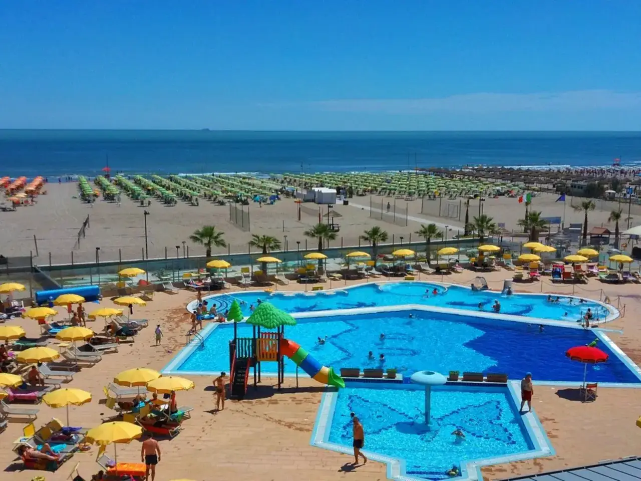 Sonnige Tage am Strand von Rosapineta Sud. Ein Pool mit Rutsche lädt zum Planschen ein, während am Meer Liegen und Sonnenschirme auf Besucher warten.