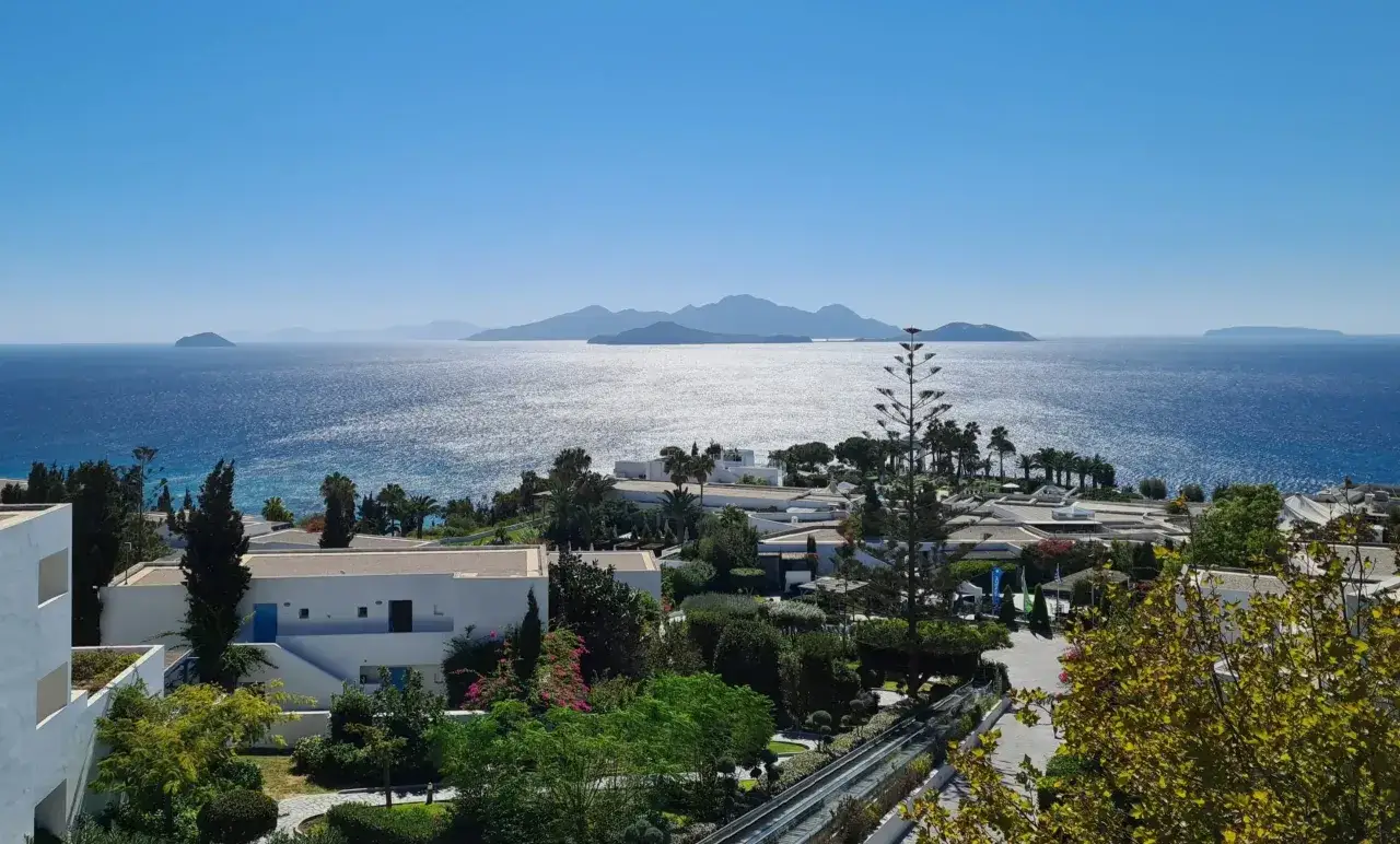 Blick auf eine Küstenanlage mit weißen Gebäuden und üppiger Vegetation, im Hintergrund das glitzernde Meer und Inseln. Ein Ort wie Robinson Daidalos, wo man sich wie ein Entdecker fühlt.