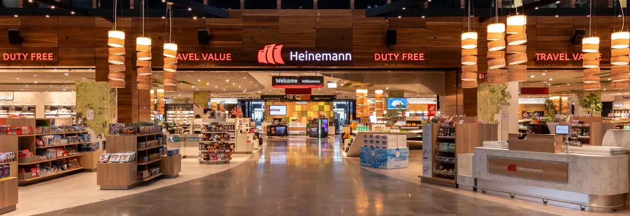 Der Eingangsbereich eines modernen duty free Shops mit dem Namen Heinemann. Regale voller Waren und ein Willkommensschild laden zum Einkaufen ein.