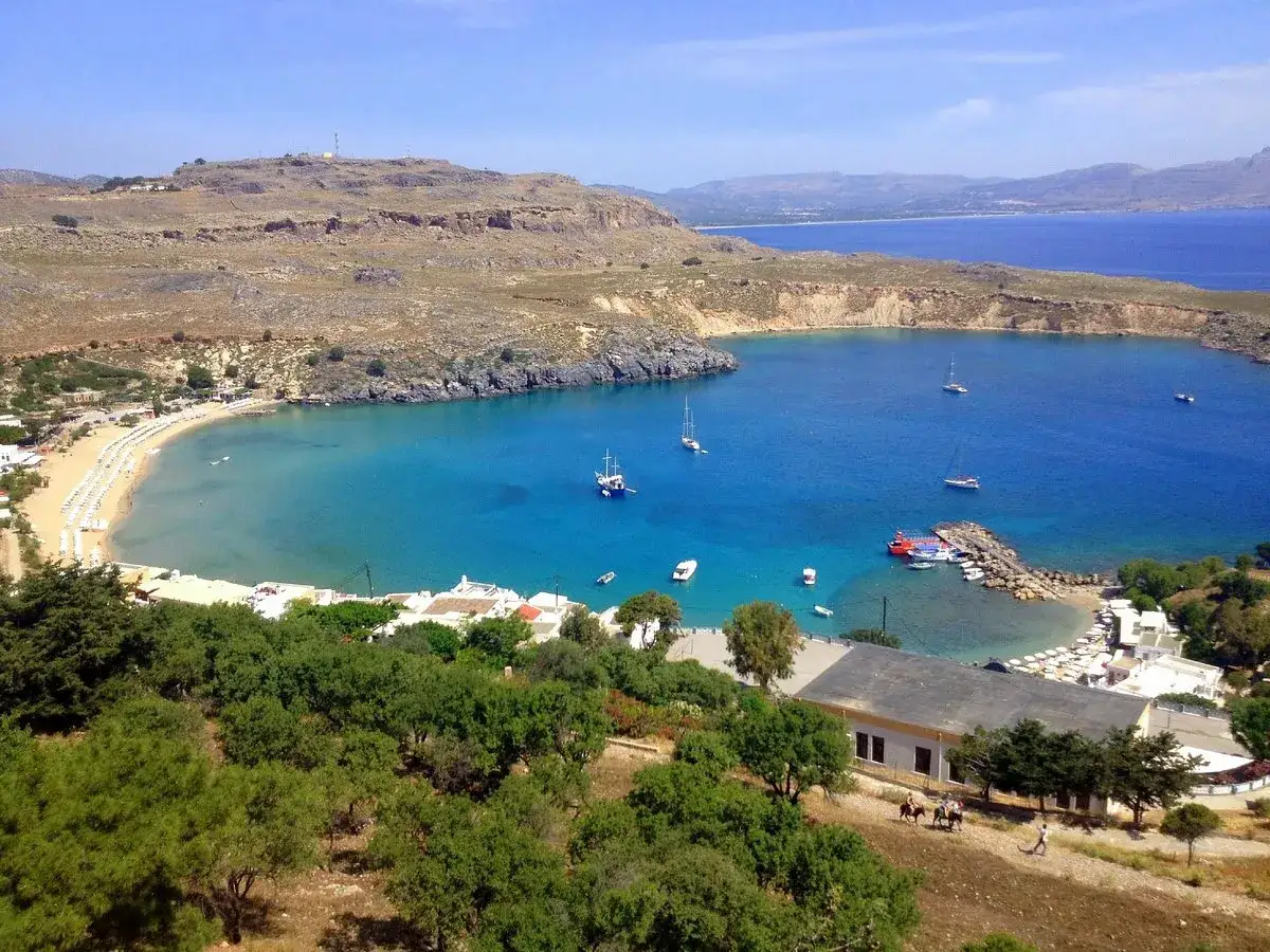 Traumhafter Blick auf Lindos, Rhodos: Türkisfarbenes Meer, weiße Liegen am Strand, Boote im Hafen und grüne Hügel.
