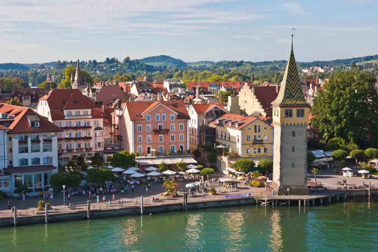 Bunte Häuserfront am Seeufer, ein markanter Turm und Menschen genießen die Sonne. Perfekt für eine TUI Bahnreise.