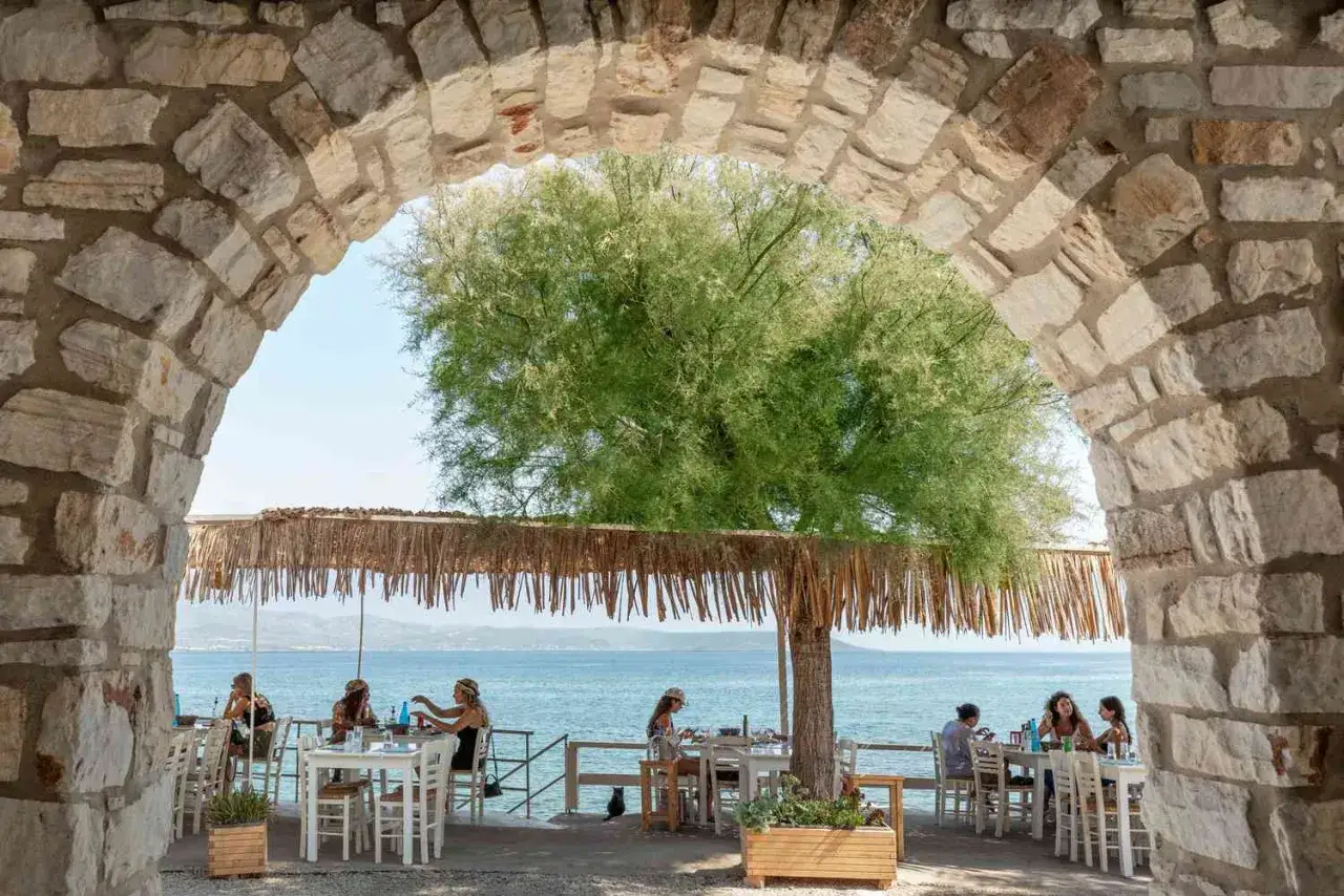 Urige Steineinfassung mit Blick auf ein Restaurant am Meer auf Paros, Griechenland. Gäste genießen das Essen unter einem Baum.