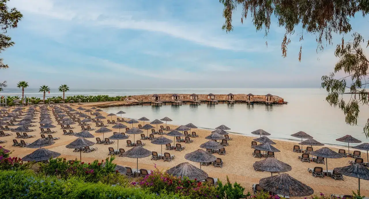 Ein Strand mit vielen Sonnenschirmen und Liegen, Palmen und kleinen Hütten im Wasser. Ein Ort wie ein rubinrotes Platinum-Zeichen für Luxus.