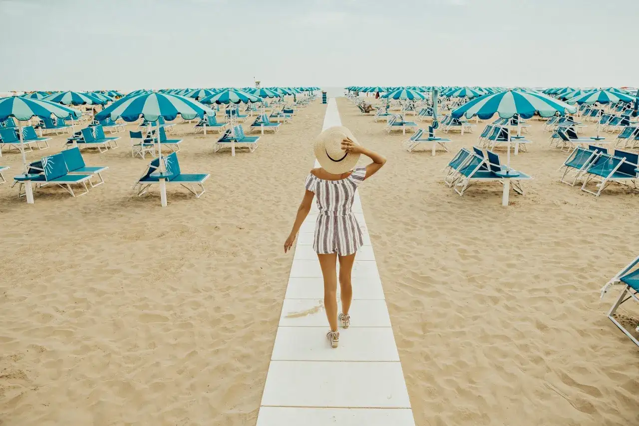 Frau mit Hut geht auf einem Steg zu den blauen Liegen und Sonnenschirmen am Strand von Rimini. Perfekter Urlaub!