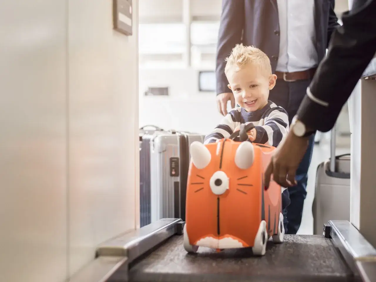 Kleiner Junge lacht, während sein orangefarbener Koffer in Form eines Tigers auf dem Gepäckband am Flughafen steht.