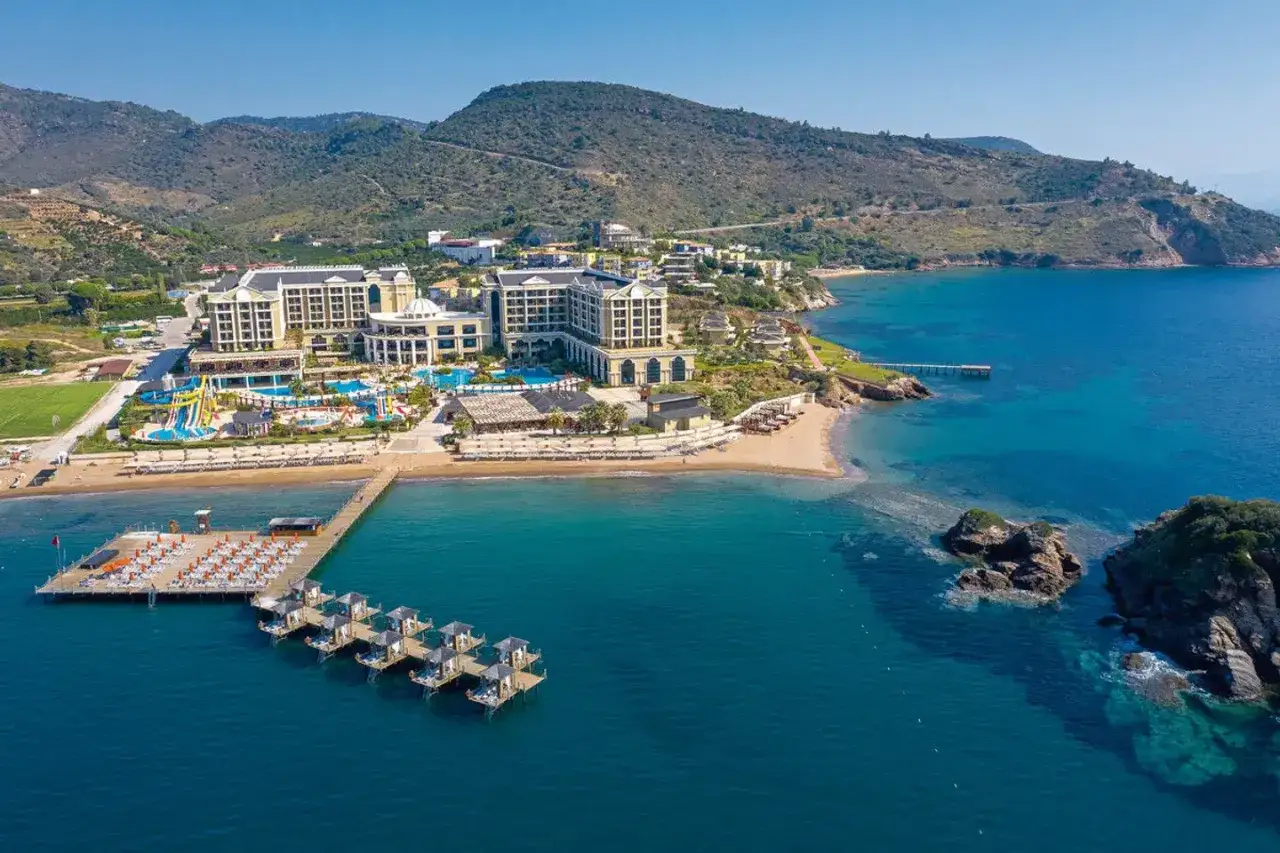 Suni's Efes Royal Palace: Ein luxuriöses Resort mit Pools, Wasserrutschen und einem Steg am türkisfarbenen Meer, eingebettet in eine hügelige Küstenlandschaft.