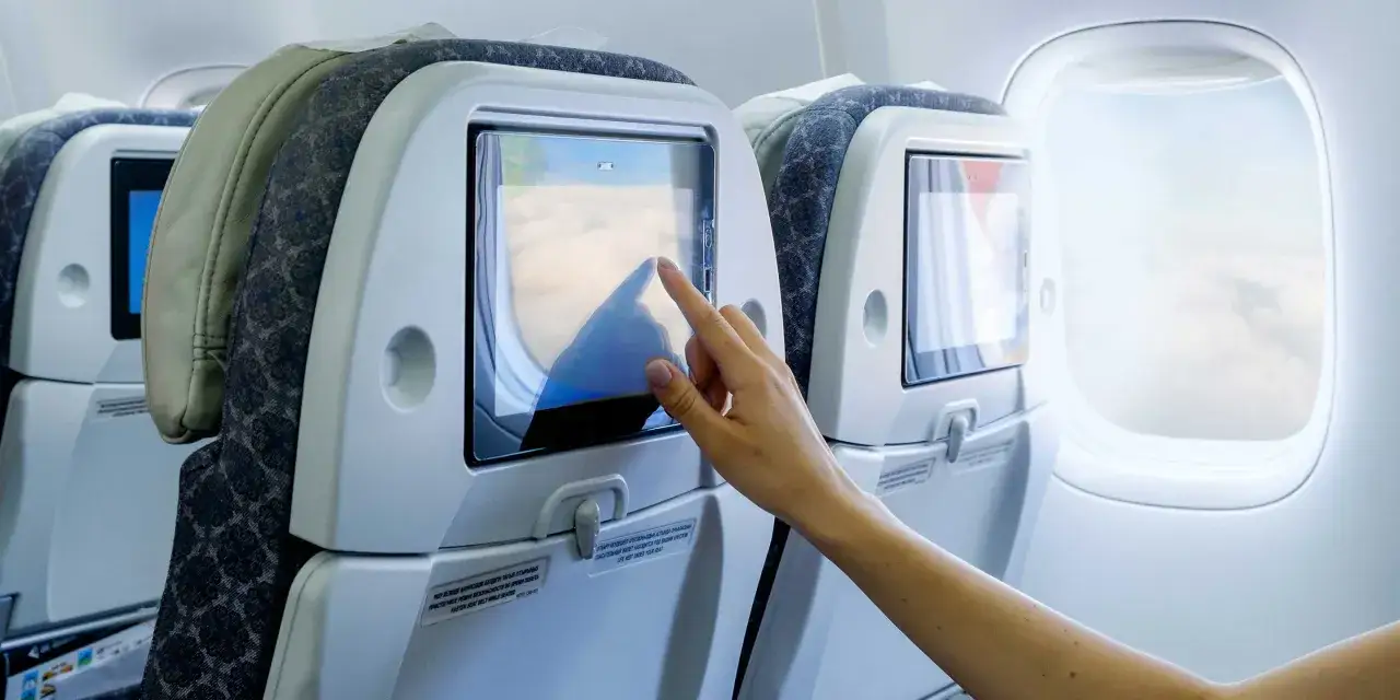 Eine Hand tippt auf den Bildschirm eines Unterhaltungssystems im Flugzeug. So kann man im Flugzeug Filme schauen oder Spiele spielen.
