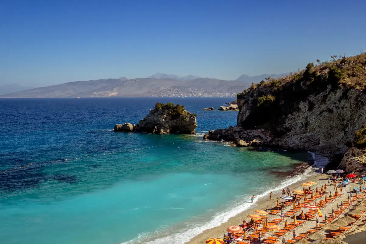 Türkisblaues Wasser trifft auf Kiesstrand mit vielen Liegen und Sonnenschirmen. Ein Felsen ragt aus dem Meer, im Hintergrund Berge. Ein Traumhafter tirana strand.