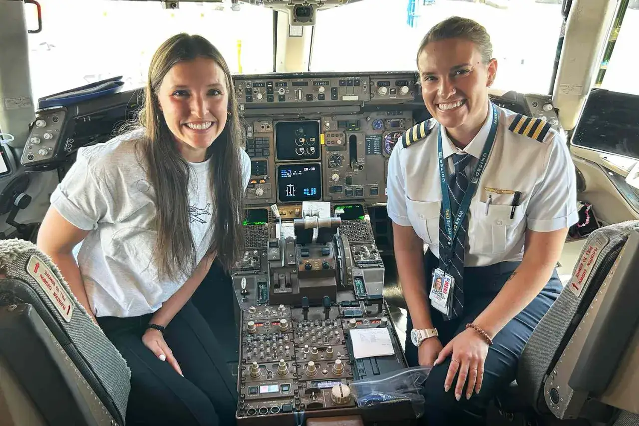 Zwei Frauen im Cockpit eines Flugzeugs. Eine trägt eine Uniform, die auf eine Pilot:in hindeutet, und lächelt. Die andere, in einem T-Shirt, sitzt daneben. Der Pilot:in Ansage Text ist auf dem Ausweis zu sehen.