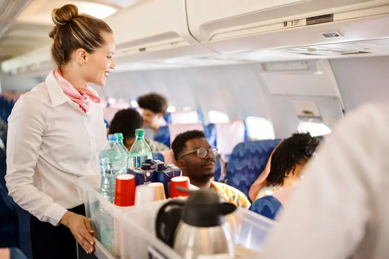 Flugbegleiterin mit einem Tablett voller Getränke und Tassen, lächelnd zu Passagieren im Flugzeug.