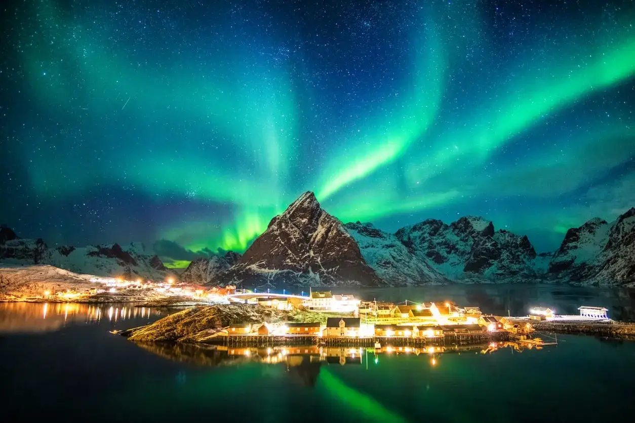 Ein Traum von einem Skandinavien Urlaub: Nordlichter tanzen über schneebedeckten Bergen und einem malerischen Dorf am Wasser.