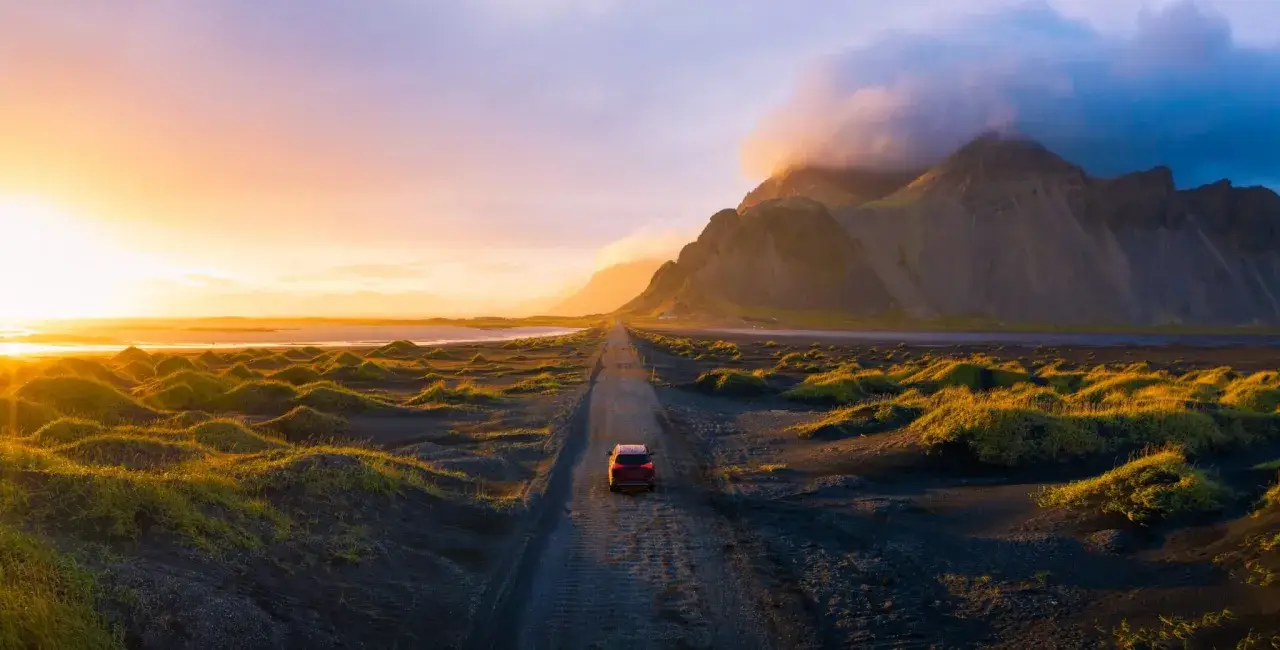 Ein rotes Auto fährt auf einer Schotterstraße durch eine isländische Landschaft bei Sonnenuntergang. Die beste Reisezeit Island für solche Ausblicke ist oft der Sommer.