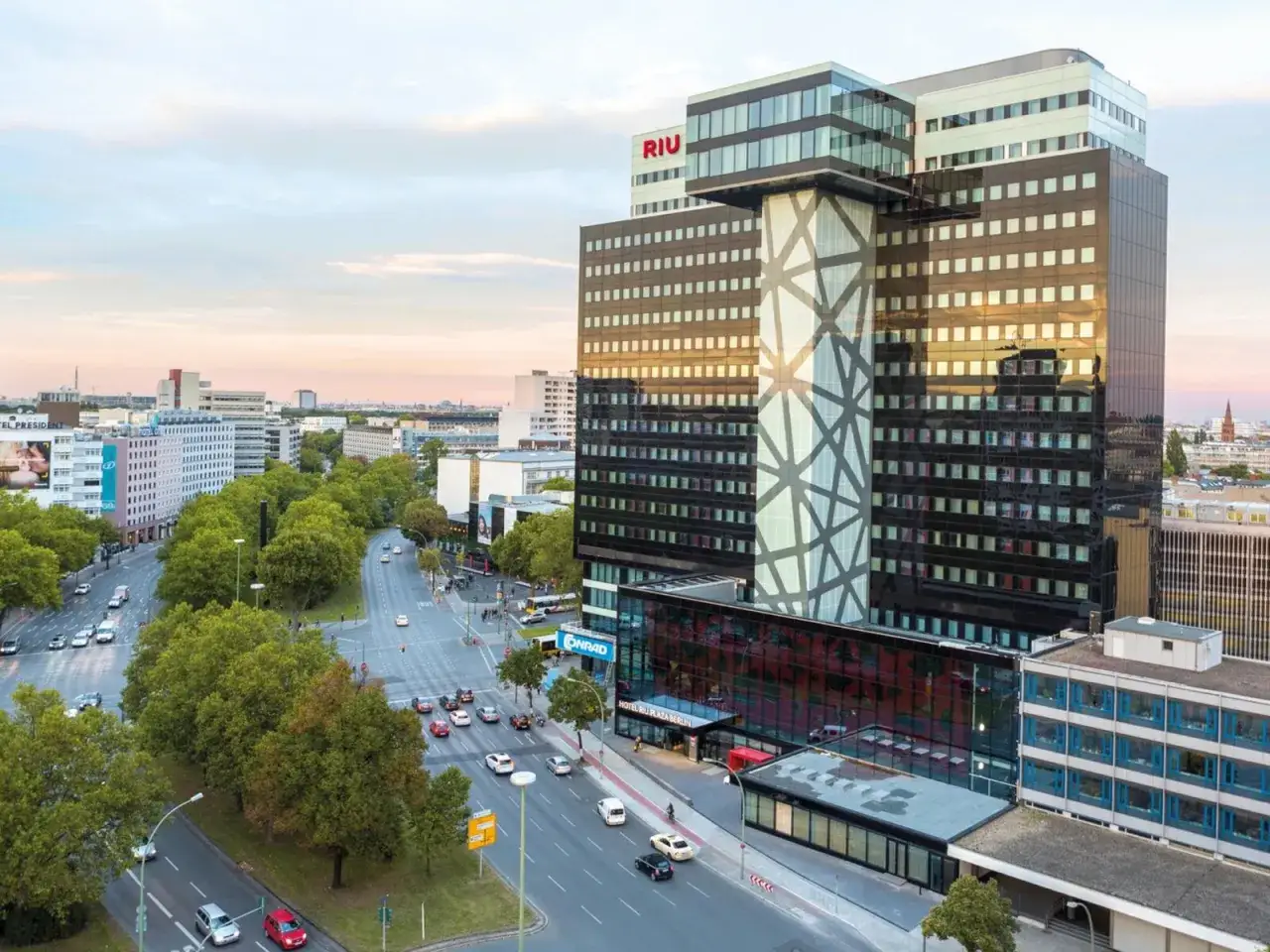 Das Hotel Riu Plaza Berlin, ein markantes Geb&auml;ude mit Glasfassade und geometrischem Muster, ragt &uuml;ber eine belebte Stra&szlig;e mit B&auml;umen und Verkehr.