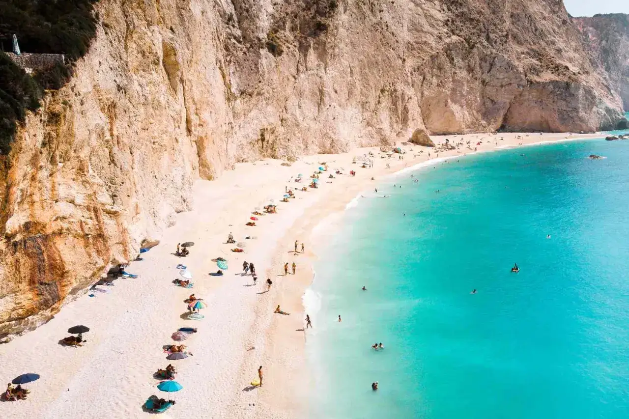 Traumhafter Urlaub Griechenland: Weiße Sandbucht mit türkisfarbenem Meer, umrahmt von steilen Felsen. Menschen entspannen unter bunten Sonnenschirmen.