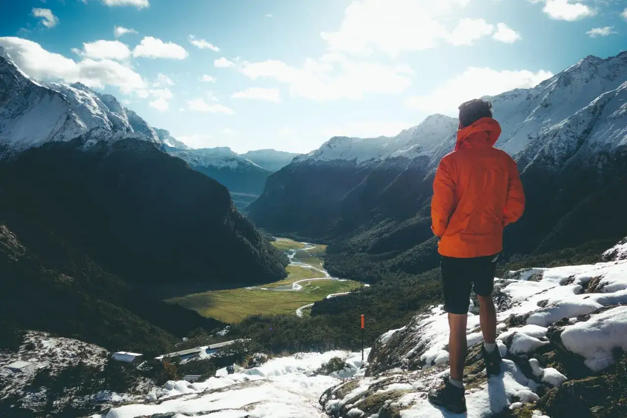 Wanderer in orange Jacke blickt auf schneebedeckte Berge und ein grünes Tal in Neuseeland. Perfekt für die Packliste Neuseeland.