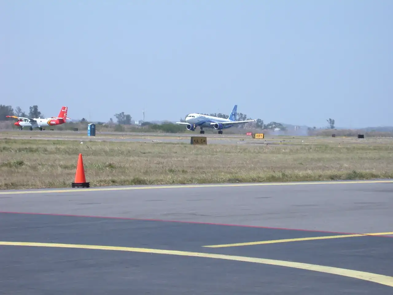 Ein blau-weißes Flugzeug nähert sich der Landebahn. Ein kleineres Flugzeug mit rotem Heck steht daneben. So landet ein Flugzeug.