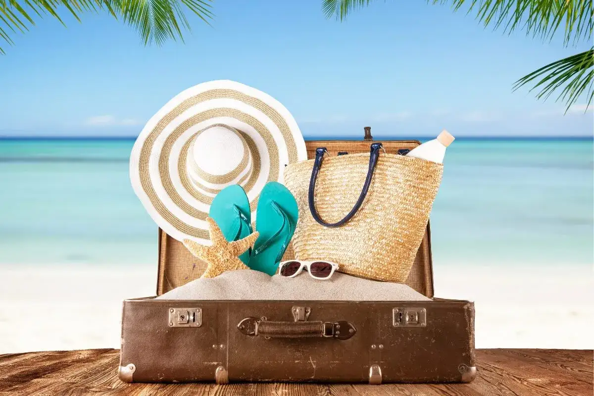 Packliste Mauritius: Koffer mit Hut, Flip-Flops, Sonnenbrille, Strandtasche und Sonnencreme am Strand.