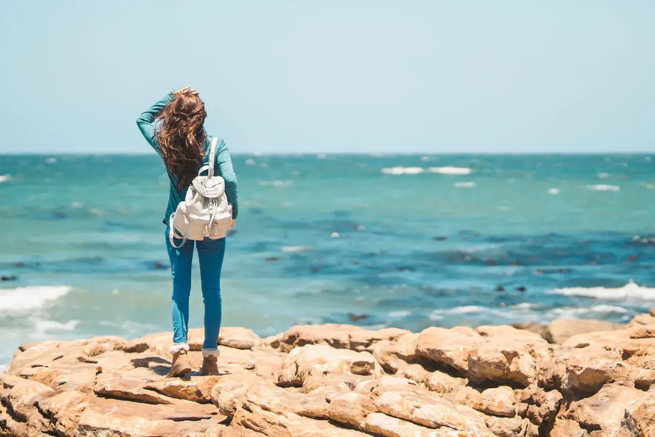 Frau mit Rucksack steht auf Felsen am Meer. Perfekt für die Packliste Malta: leichte Kleidung, Sonnencreme und gute Schuhe für Erkundungen.
