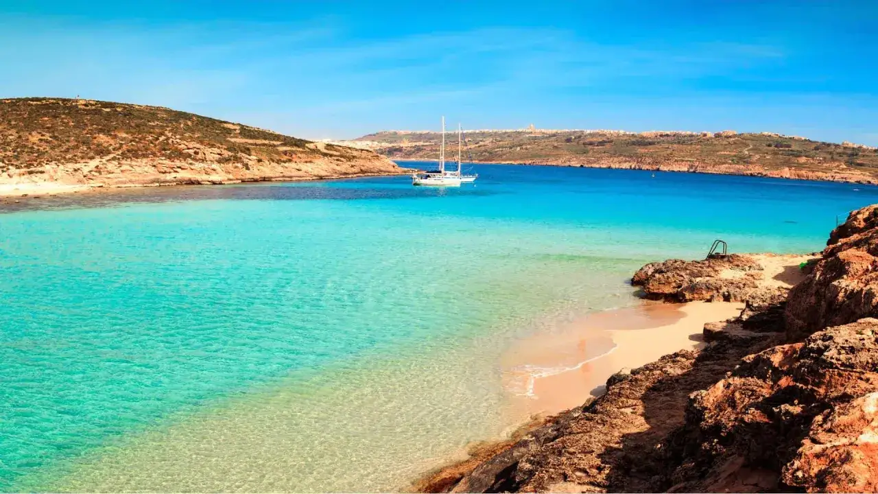 Traumhafter Urlaub auf Malta: Türkises Wasser, Sandstrand und ein Segelboot in einer malerischen Bucht.