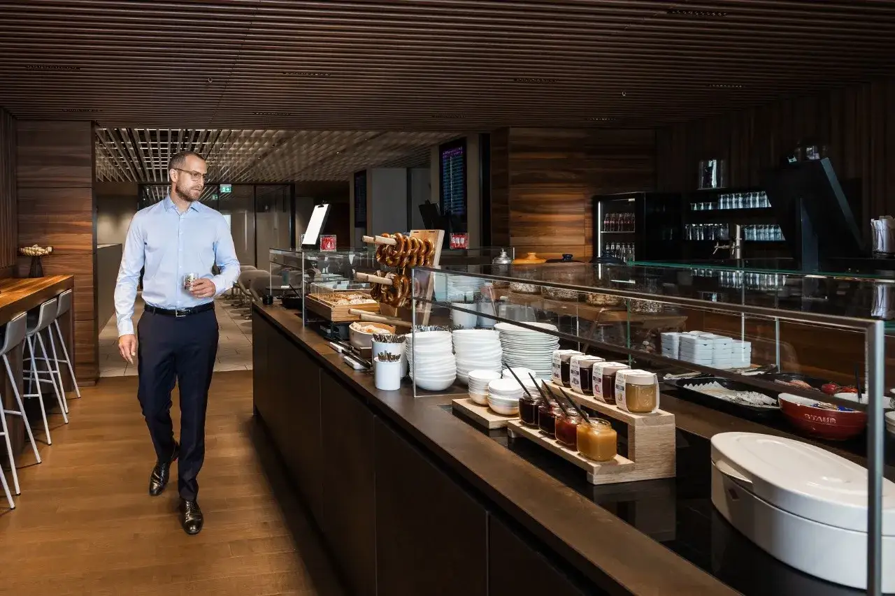 Mann in einem Anzug geht durch die Priority Pass Lounge Berlin, vorbei an einem Buffet mit Brezeln, Marmeladen und Geschirr.