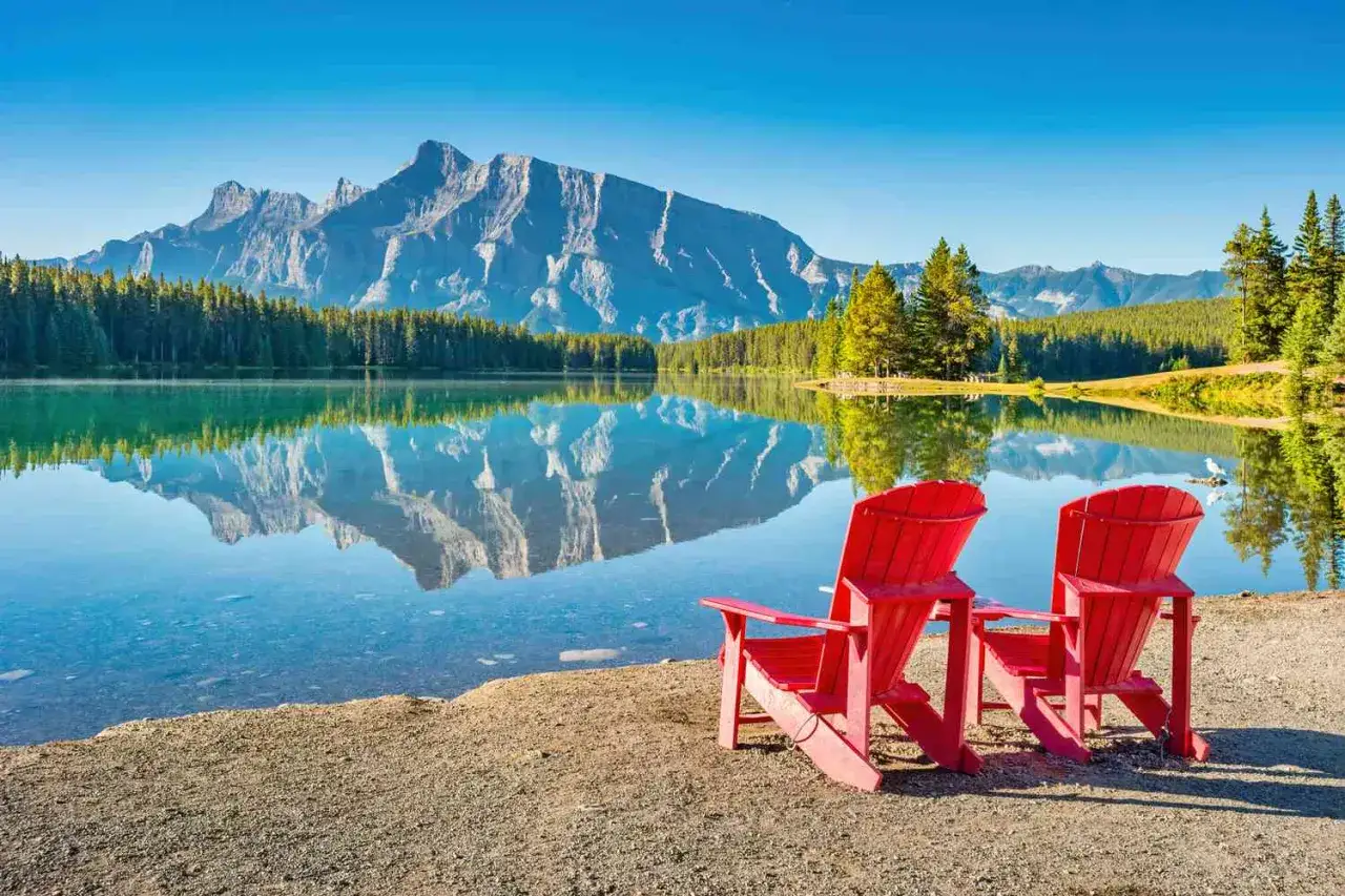 Zwei rote Adirondack-Stühle am Ufer eines ruhigen Sees mit Bergspiegelung. Perfekt für einen Urlaub in Kanada.