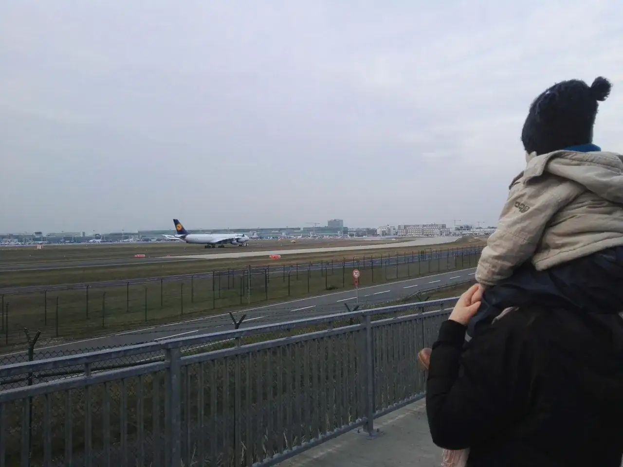 Kind auf den Schultern der Mutter genie&szlig;t das Plane Spotting am Flughafen. Ein Lufthansa-Flugzeug rollt auf der Startbahn.
