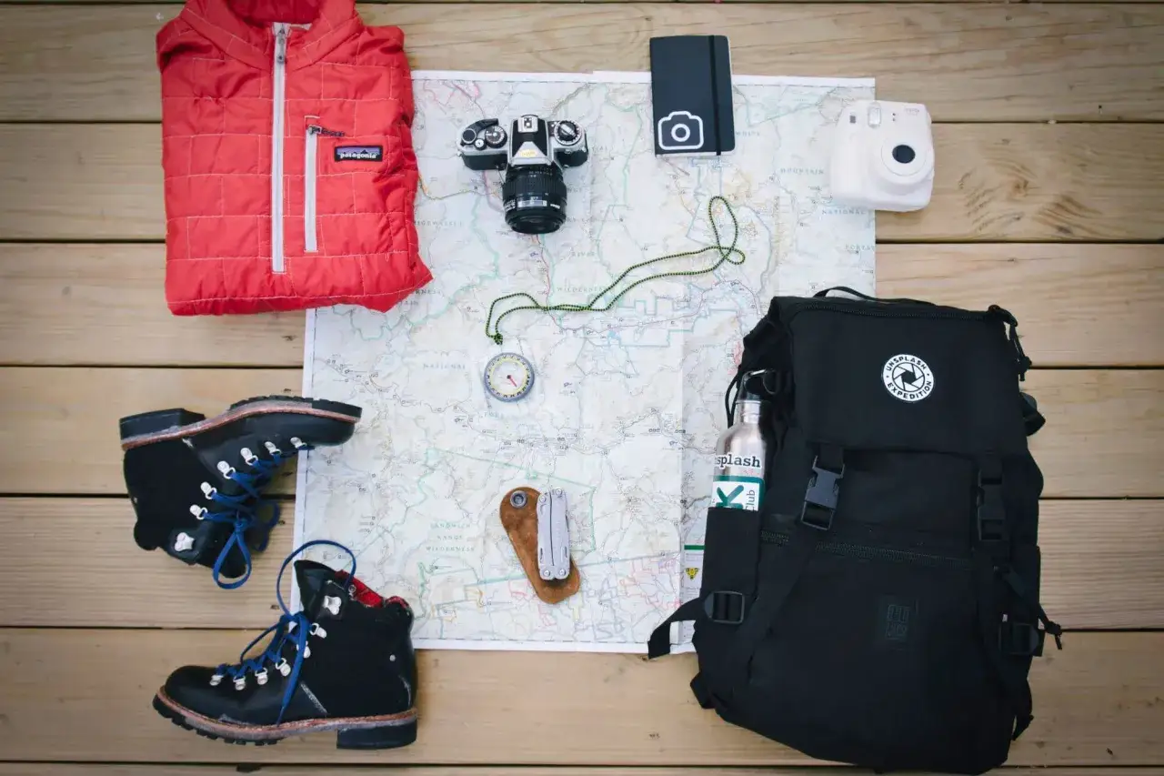 Packliste Kanada: Wanderschuhe, rote Daunenjacke, Kamera, Karte, Kompass, Multitool, Rucksack und Wasserflasche für das Abenteuer.