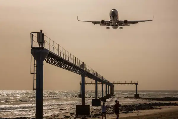 Zwei Personen genießen das Plane Spotting am Strand, während ein Flugzeug über eine Seebrücke hinwegfliegt.