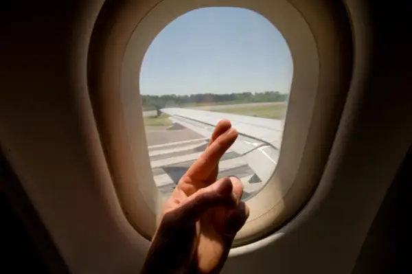 Hand mit überkreuzten Fingern vor Flugzeugfenster. Ein Zeichen der Hoffnung für alle Flugzeug-Passagiere.