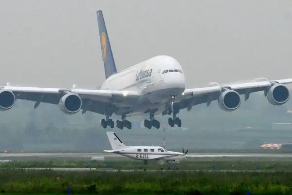Ein riesiger A380 von Lufthansa landet, sein enormes Gewicht wird durch die vier Triebwerke und das Fahrwerk sichtbar. Ein kleines Propellerflugzeug wartet daneben.